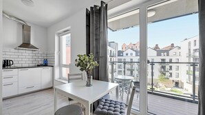 Cocina moderna con armarios blancos, campana extractora de acero inoxidable y un comedor con mesa blanca y sillas grises acolchadas. Un jarrón con plantas verdes está sobre la mesa.