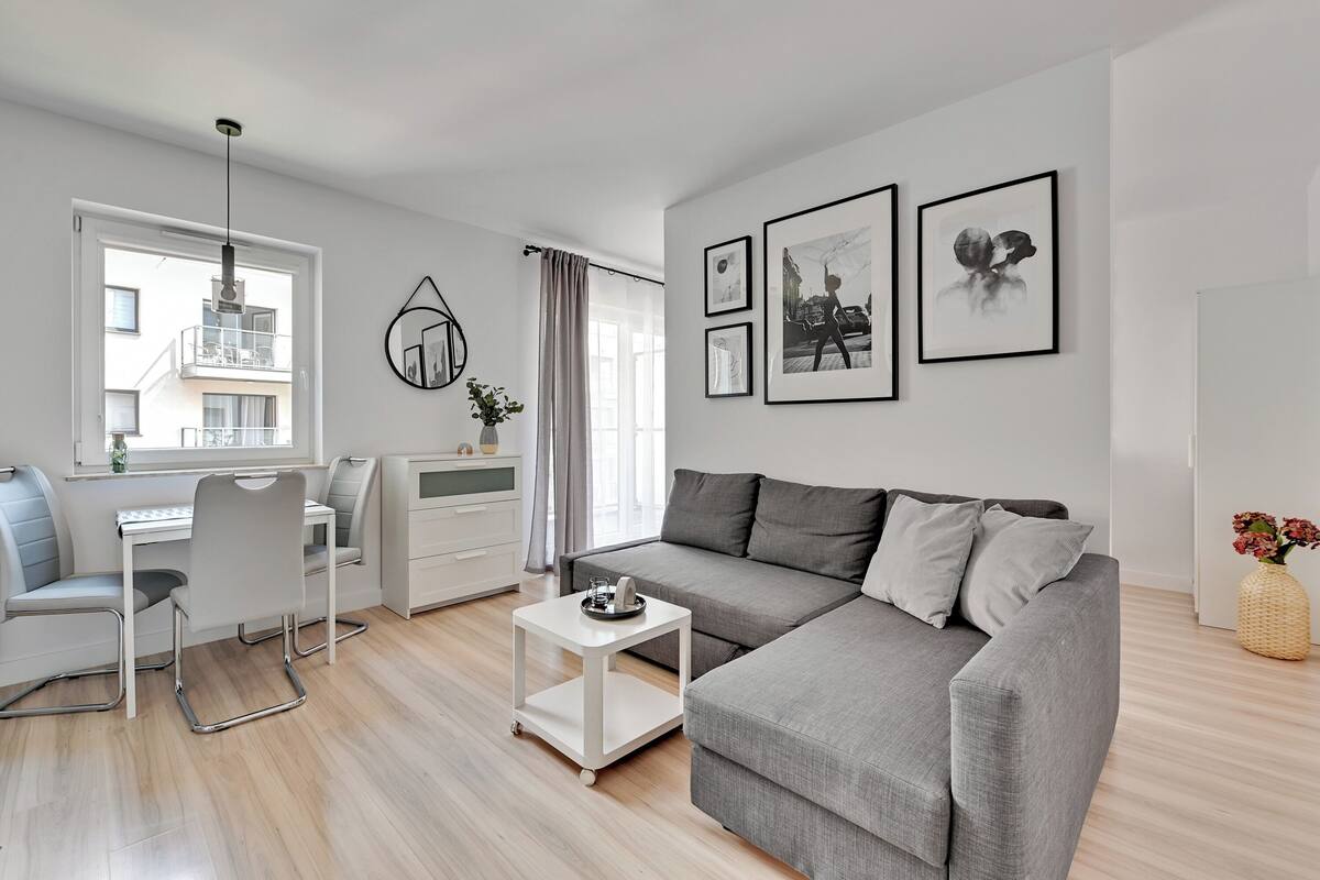 A modern living room with a grey sofa, a white coffee table, a desk with a chair, and a window with curtains. There are framed pictures on the wall.