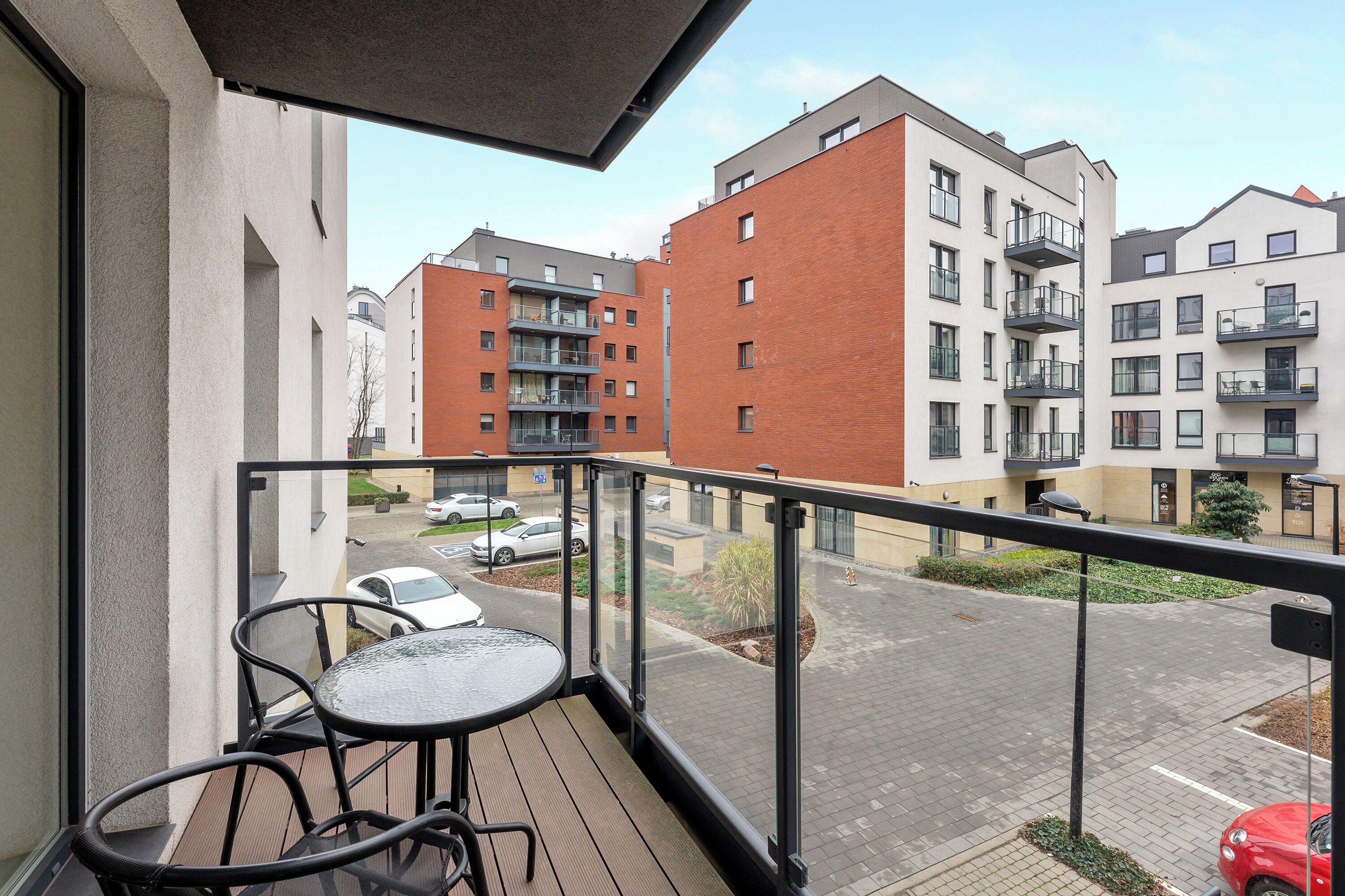 A balcony with a glass railing overlooking a parking area and adjacent buildings.