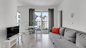 Una sala de estar moderna con una televisión de pantalla plana sobre un soporte blanco, un sofá gris con un cojín rojo, una mesa de comedor con sillas y vistas a edificios a través de amplias ventanas.