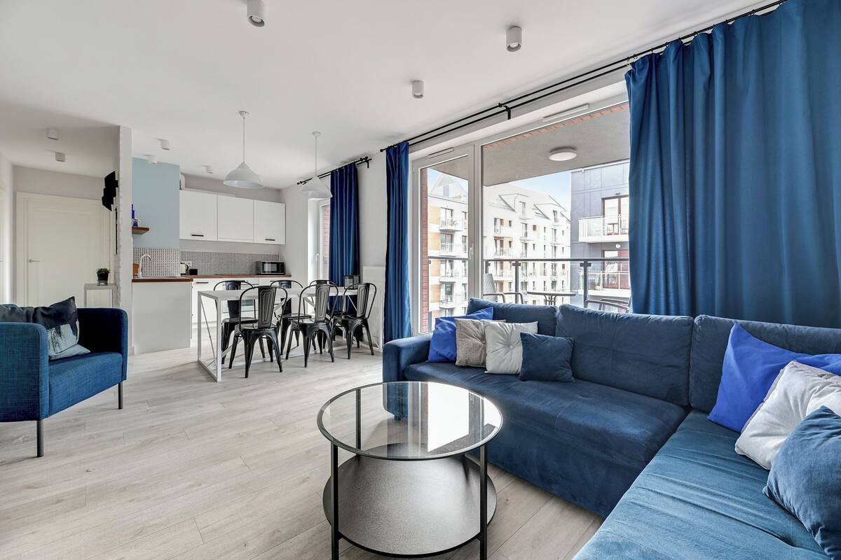 A modern living room with a blue sofa, a glass coffee table, and a view of a cityscape through large windows.