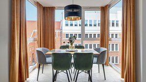Un comedor con una mesa y sillas, un jarrón con flores, y vistas a edificios a través de amplios ventanales.