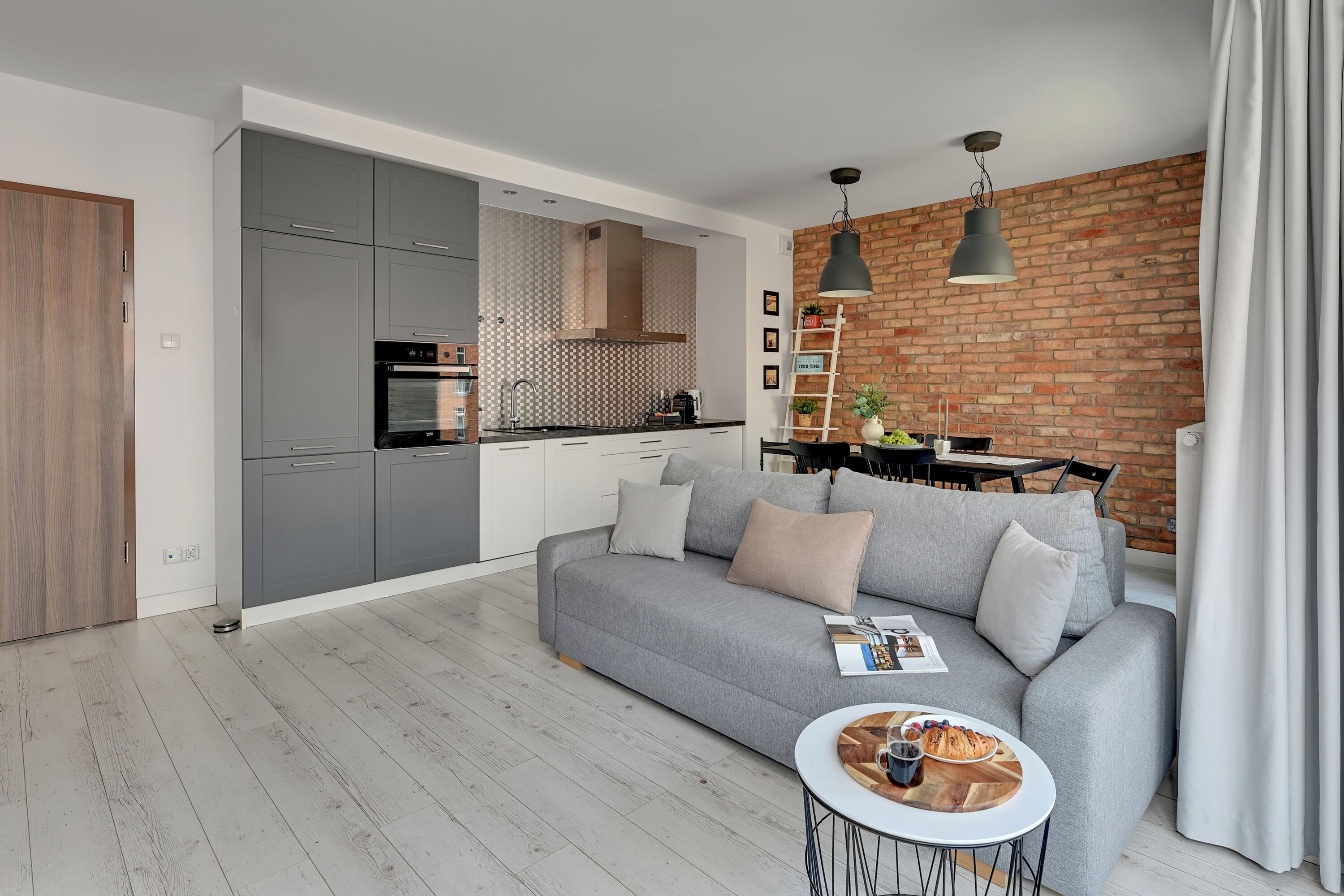 A modern living room with a grey sofa, a coffee table, and a dining area with a brick wall and pendant lighting.