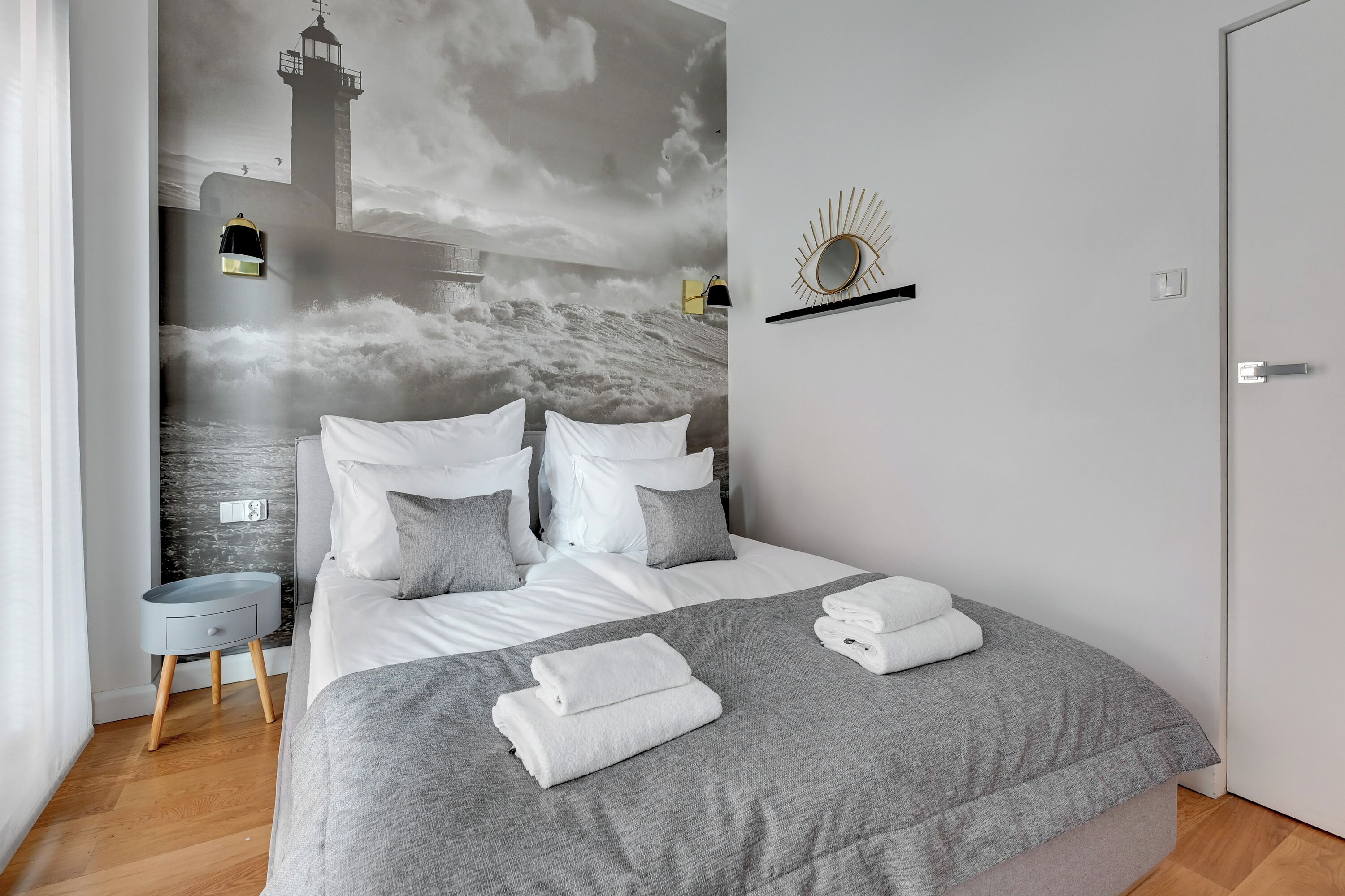 A modern bedroom with a large bed, grey bedding, and a mural of a lighthouse on the wall.