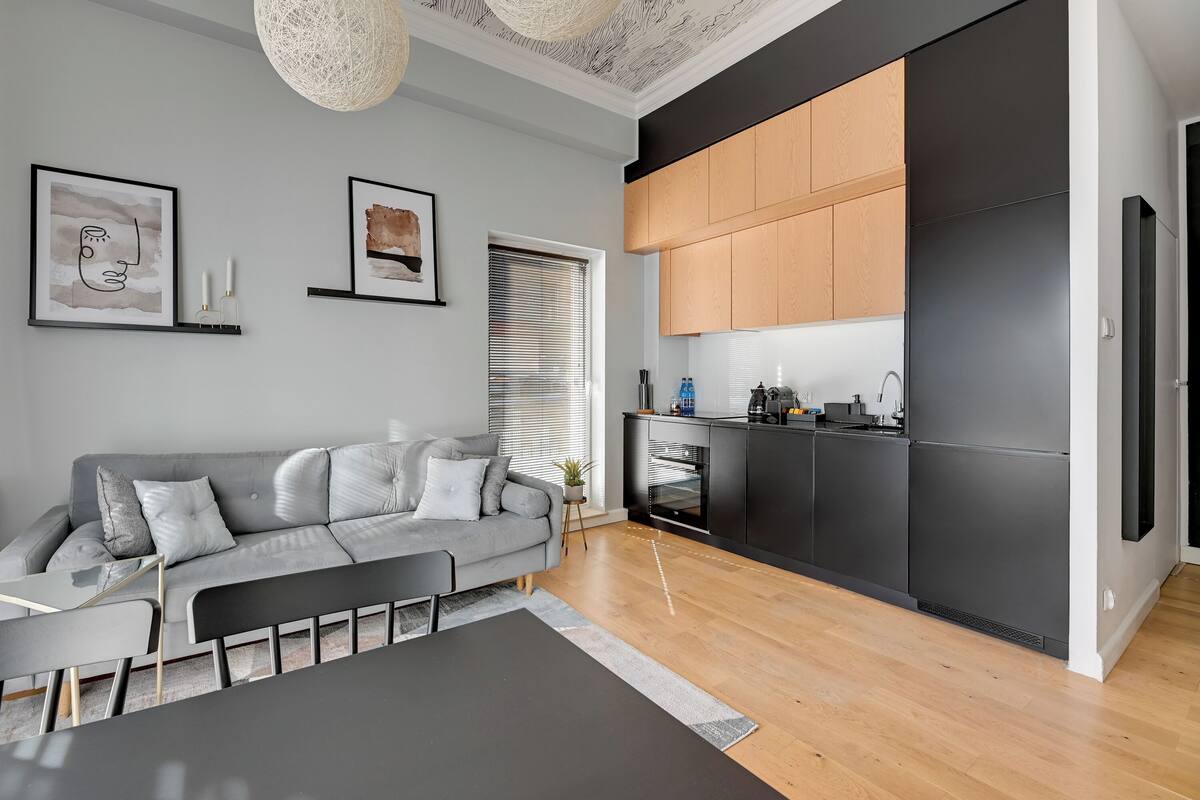 A modern living room with a grey sofa, a dining table, and a kitchen area with black cabinets and wooden countertops.
