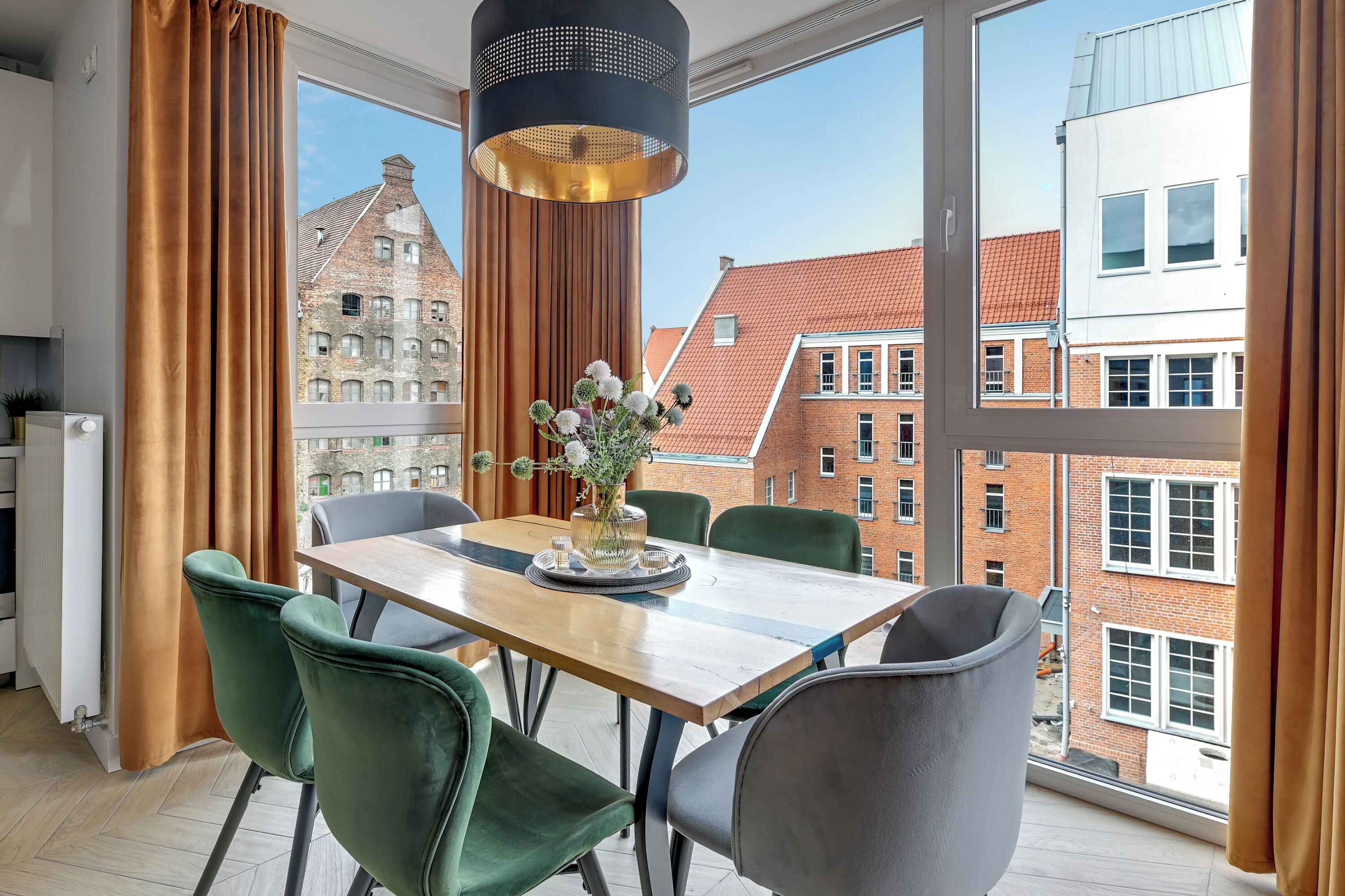 Ein moderner Essbereich mit Holztisch, grünen und grauen Stühlen, einer Blumenvase und einer schwarzen Pendelleuchte.