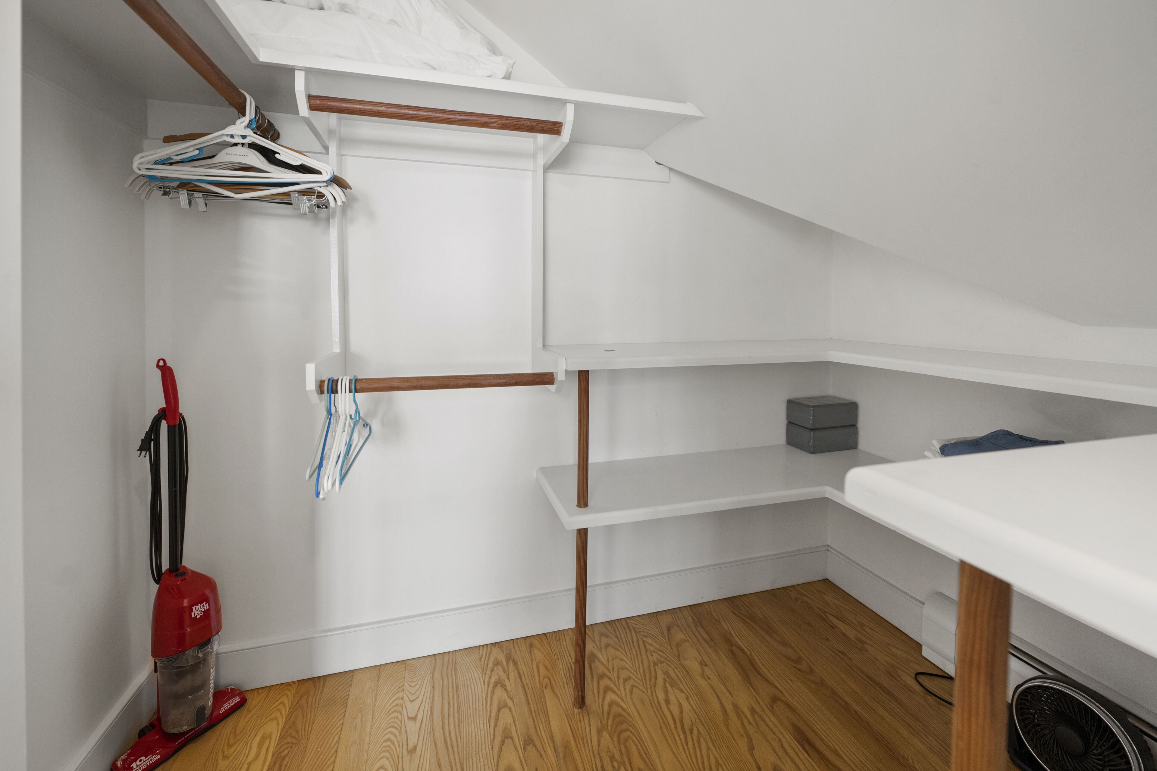 Un coin de pièce avec un mur blanc, un plancher en bois, une étagère blanche, un aspirateur rouge et une table blanche.