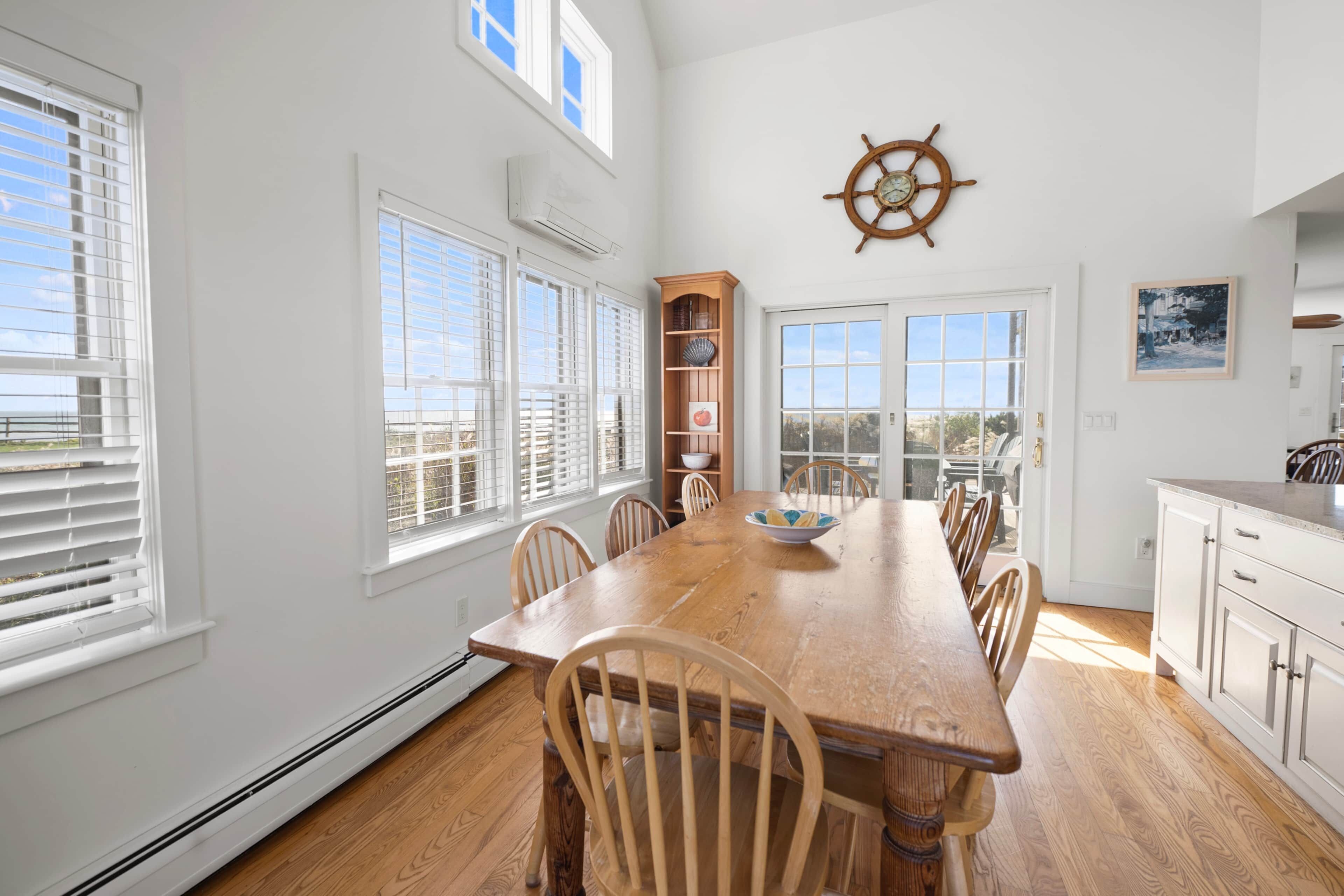 A dining area with a wooden table and chairs, a ship's wheel decoration, and a view through the glass door.