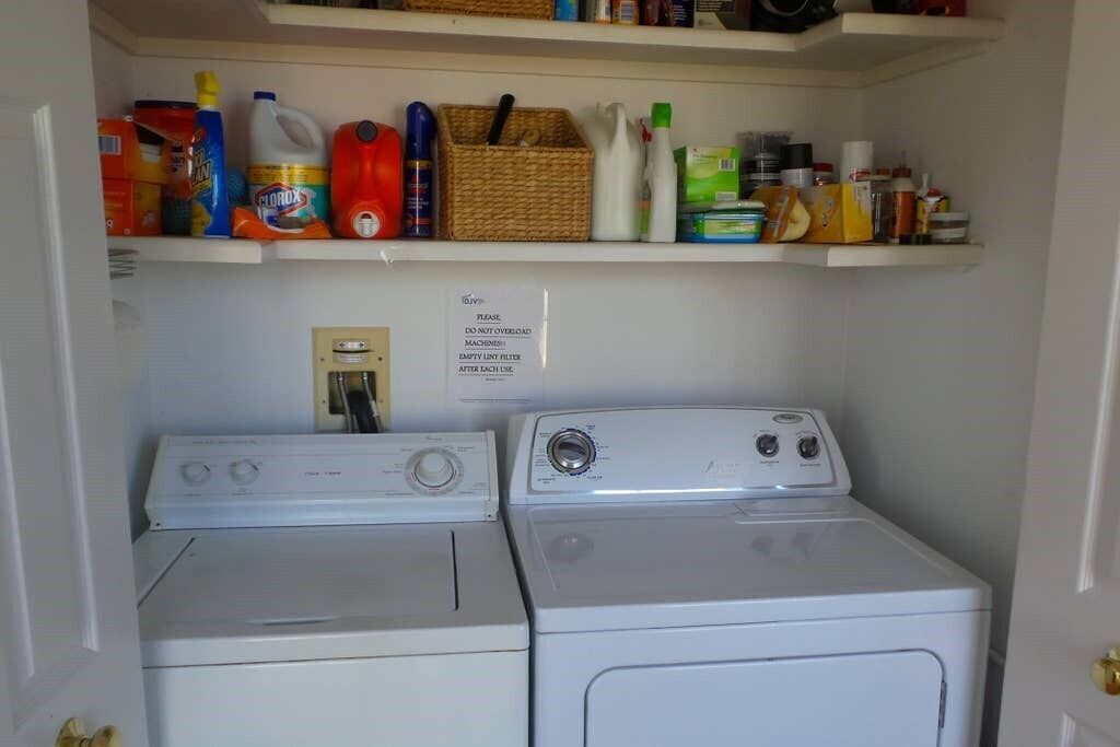 A laundry area with two washers and dryers, a shelf above with cleaning supplies, and a sign instructing not to overload the machines.