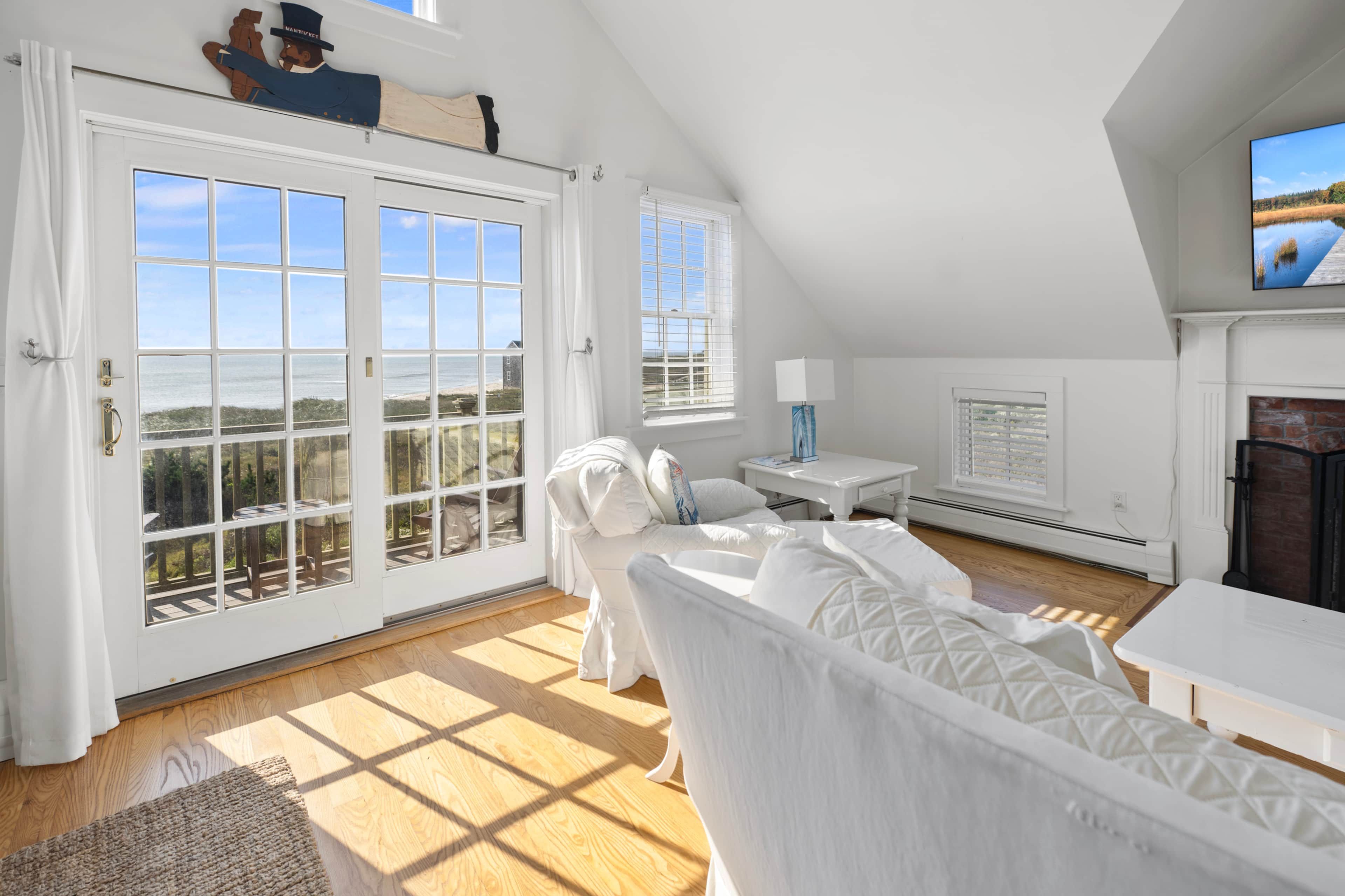 A spacious room with a large bay window overlooking a beach, a fireplace, and a cozy seating area.