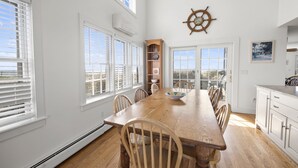 A dining area with a wooden table, chairs, a ship's wheel wall decoration, and large windows.