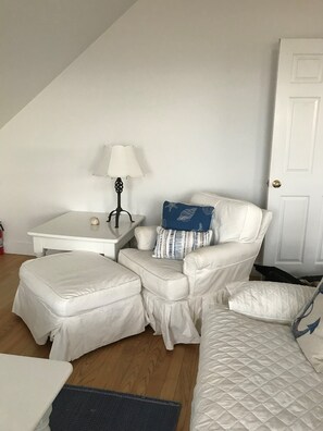 A small, white, loft-style bedroom with a single white chair, a small white table, and a lamp.