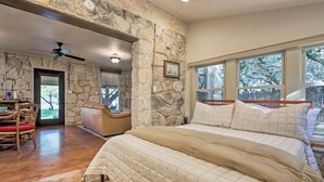 Camera da letto con parete in pietra, un letto grande con biancheria bianca, una ventola a soffitto e vista su alberi attraverso ampie finestre.
