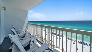Ein Balkon mit weißen Stühlen mit Blick auf einen Strand mit Sonnenschirmen und Menschen.