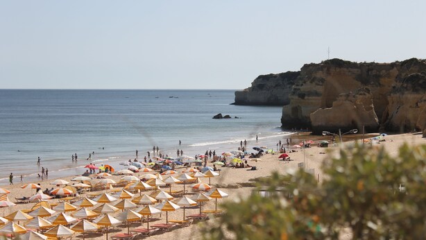 Ein Strand mit vielen Sonnenschirmen und Liegestühlen, einer felsigen Klippe und dem Meer.