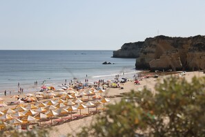 A beach with numerous umbrellas and sun loungers, a rocky cliff, and the sea. - Encarnado - 1 bedroom apartment (Armacao De Pera)