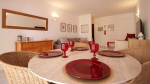 Ein Essbereich mit einem runden Tisch, gedeckt für vier Personen, umgeben von Korbstühlen. Links ist ein Holzschrank und im Hintergrund eine Couch mit Kissen zu sehen.