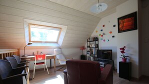 A room with a sloped ceiling, a desk with chairs, a bookshelf, a television, and a window with a view of trees.