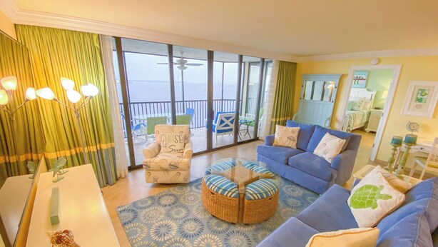 A living room with a sofa, armchair, and ottoman, a dining table, and a view of the sea through large windows. - Sanibel Harbour Resort Harbour Tower 713:   Relaxing in the tropics in this beach themed condo (Fort Myers)