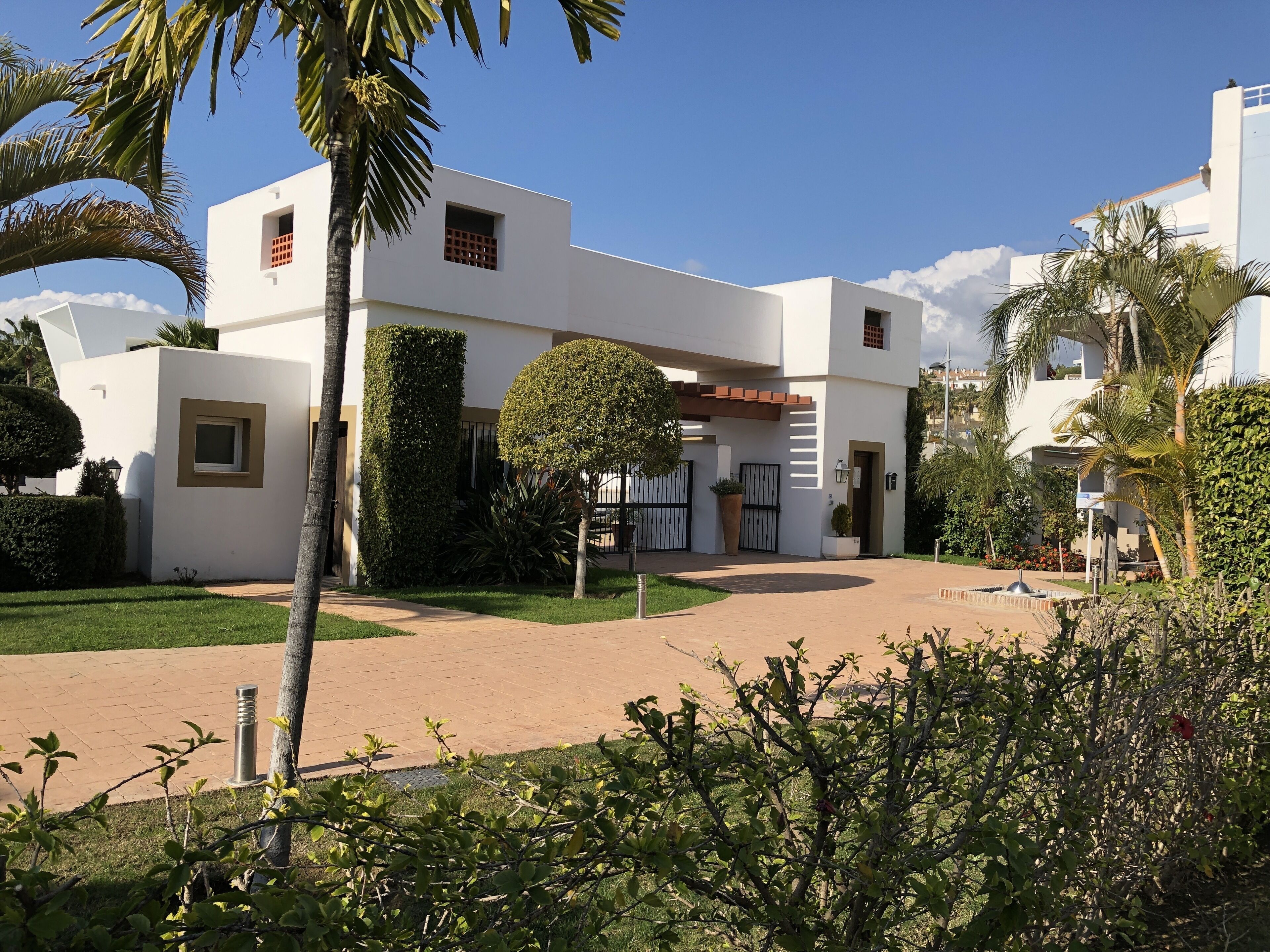 Un edificio moderno con fachada blanca, entrada central y un jardín circundante con palmeras y arbustos.