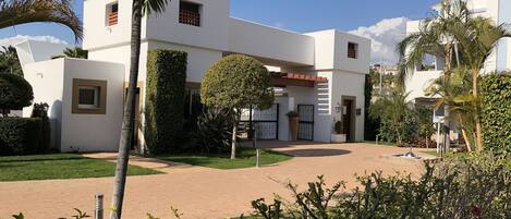 Un edificio moderno con fachada blanca, entrada central y un jardín circundante con palmeras y arbustos.