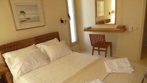 Una cama con sábanas blancas impecables, un cabecero de mimbre, una silla de madera, una mesita de noche con una lámpara y un cuadro en la pared.
