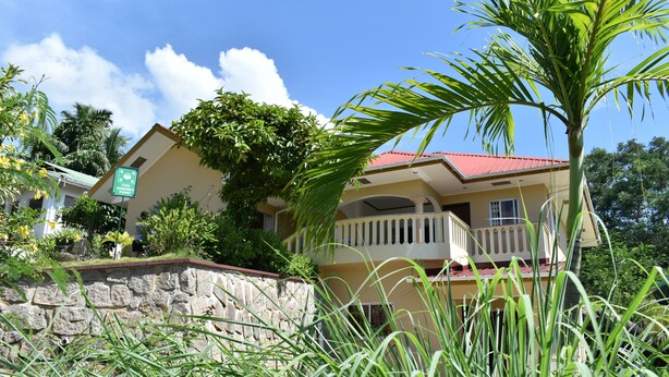 A two-story building with a stone wall in front, a red roof, and a balcony on the second floor. - Zeph Selfcatering (Mahé Island)