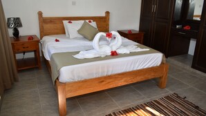 A neatly made bed with a wooden headboard, a bedside table with a lamp, and a nightstand with a vase. - Zeph Selfcatering (Mahé Island)