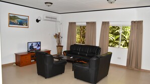 A living room with a sofa, two armchairs, a coffee table, a TV on a wooden cabinet, a potted plant, and a window with curtains. - Zeph Selfcatering (Mahé Island)