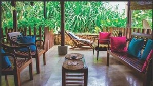 A tropical-style room with wooden furniture, a central table, and a view of lush greenery outside. - Whispering Ocean (Panadura)