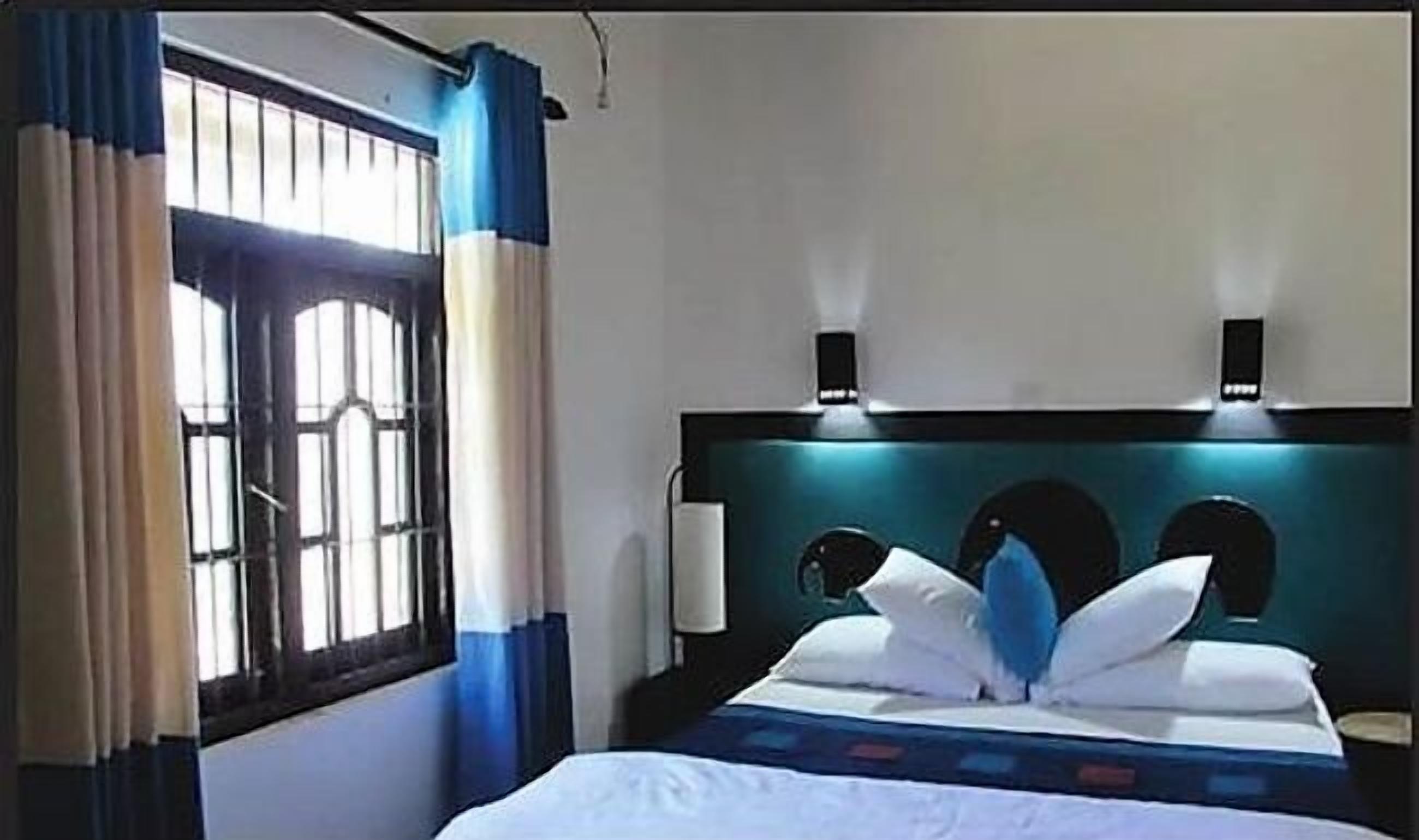 A hotel room with a bed, a window with curtains, and a headboard with wall-mounted lights.