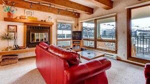 Una sala de estar acogedora con un sofá de cuero rojo, una chimenea, un televisor y grandes ventanas que ofrecen vistas al exterior.