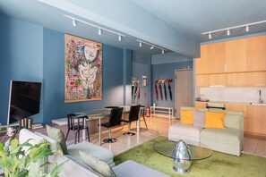 A modern living room with a blue wall, a large abstract painting, a dining area with a glass table, and a kitchen with wooden cabinets. - Central Comfy Apt with Private Balcony & Rooftop Access. CT3 (Athens)