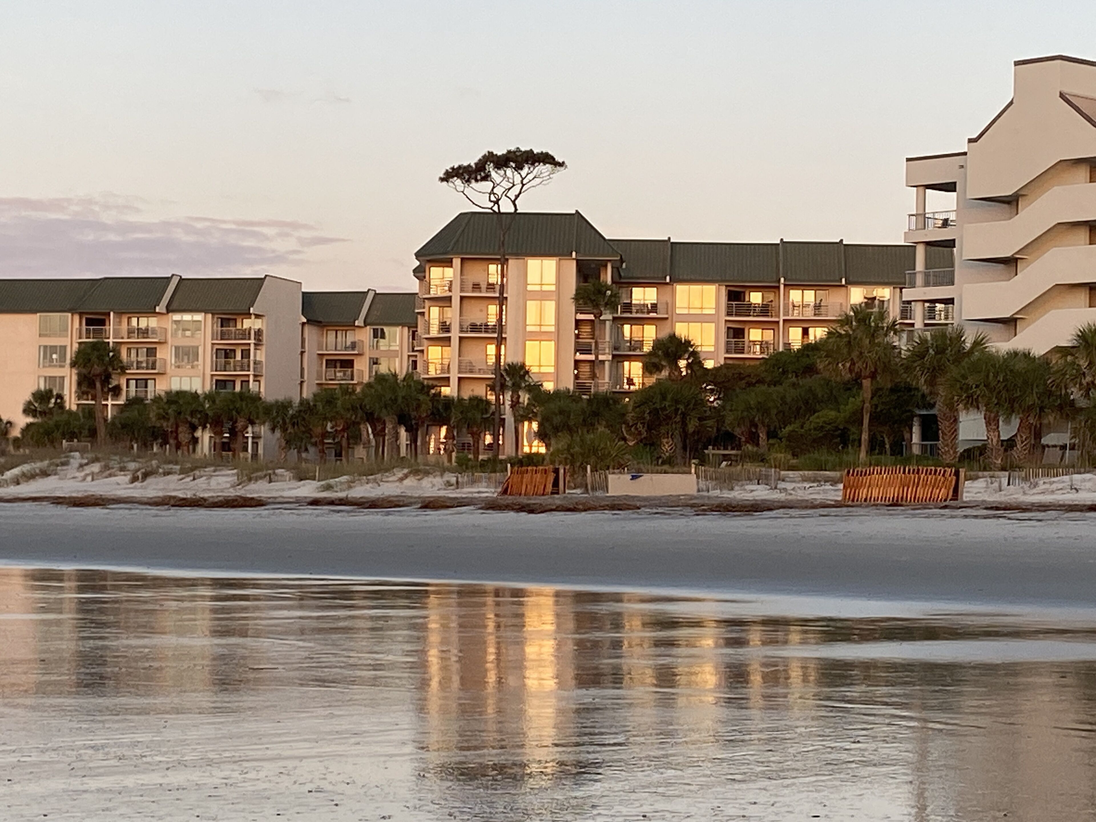 Ein Hotel direkt am Strand mit mehreren Gebäuden, einem Baum auf dem Dach des Hauptgebäudes und einem Sandstrand im Vordergrund.