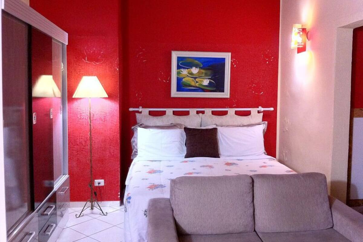 A hotel room with a red wall, a bed with white and brown pillows, a grey sofa, a painting of a landscape, and a lamp.