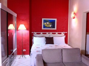A hotel room with a red wall, a bed with white and brown pillows, a grey sofa, a painting of a landscape, and a lamp. - Pousada Requinte da Mantiqueira (Piracaia)