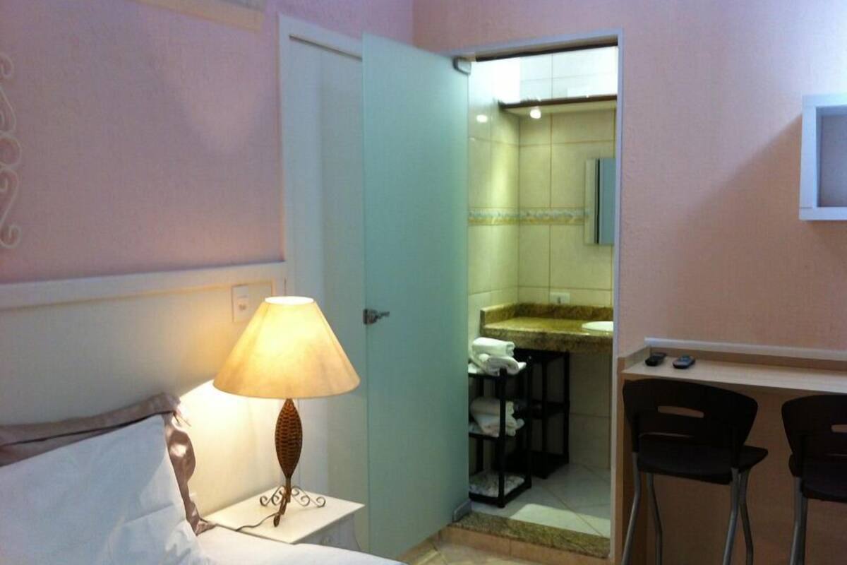 A hotel room with a bed, a nightstand with a lamp, a desk with two chairs, and a bathroom visible through an open door.