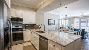A modern kitchen with white cabinets, a stainless steel refrigerator, a built-in oven, and a dishwasher. There is a large island with a sink and a pendant light. The kitchen leads to a dining area with a blue sofa and a table. - Tides at Tops`l #301- Overlooks the Gulf! (Miramar)