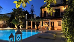 Una piscina con sillas de descanso y un comedor, rodeado de vegetación y un edificio con balcón.