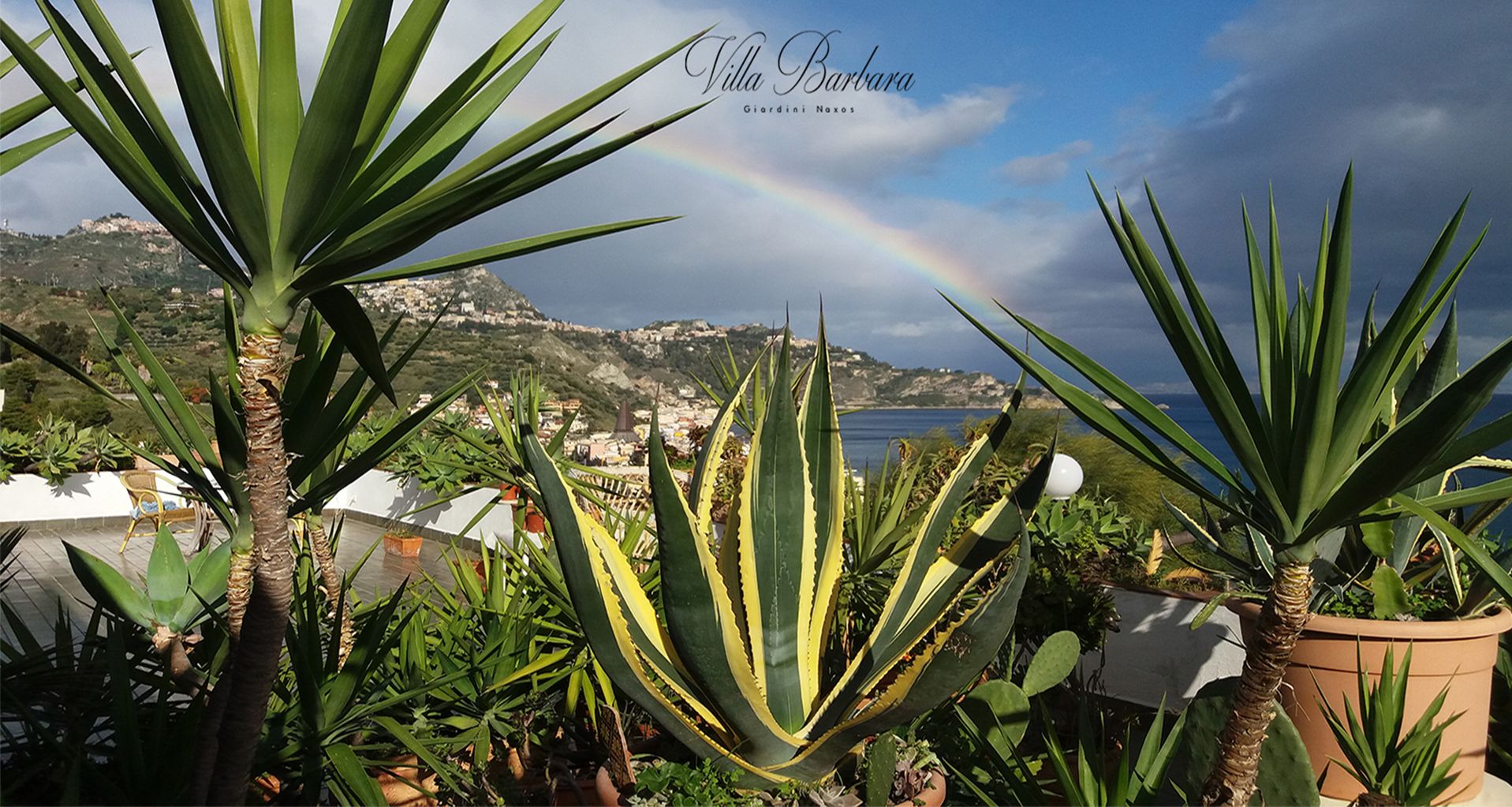 Un paisaje costero con un arcoíris, con plantas de agave y vistas al mar y las colinas.