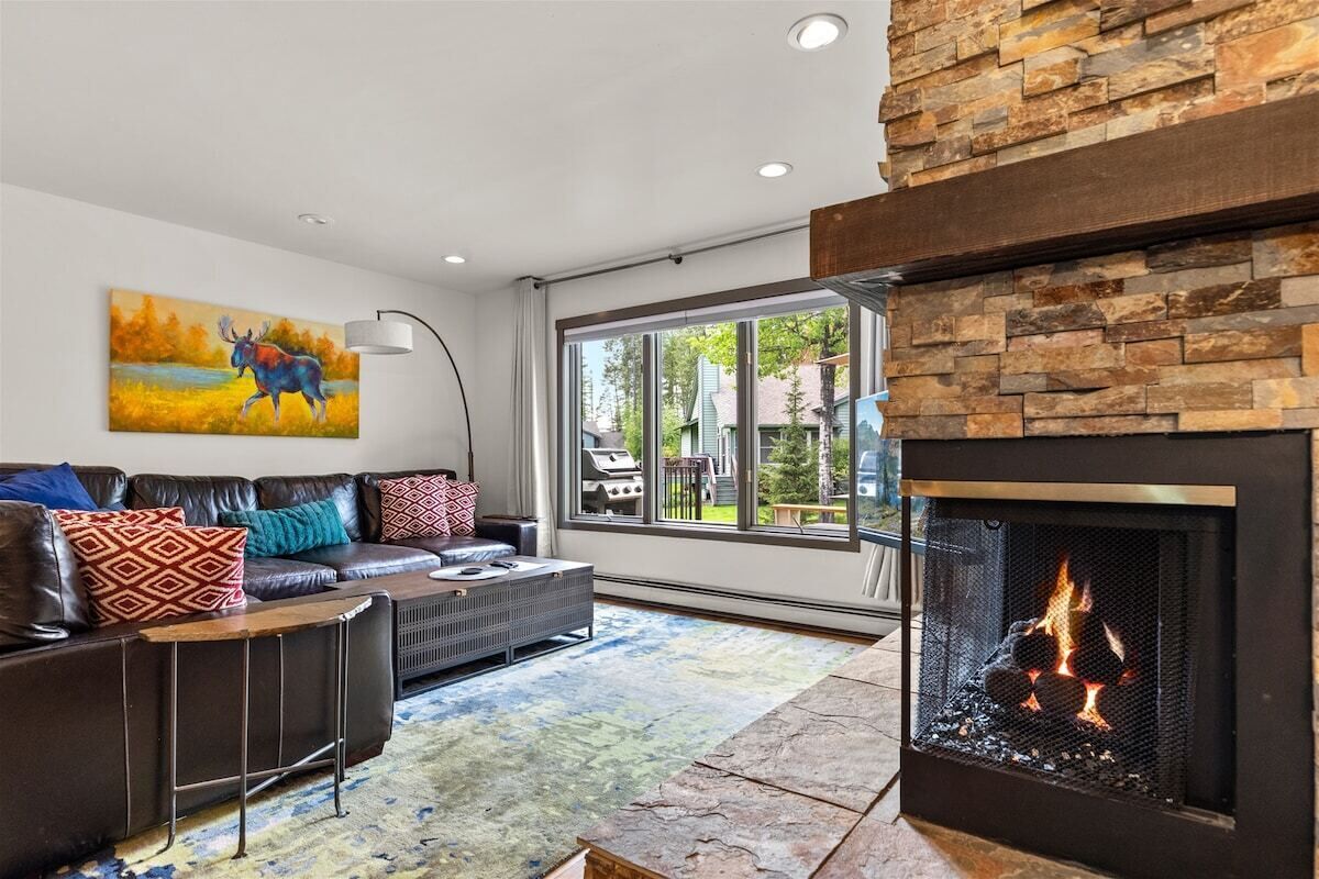 A modern living room with a fireplace, leather sofa, and a large window offering a view of the outdoors.