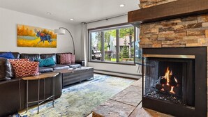 A modern living room with a fireplace, leather sofa, and a large window offering a view of the outdoors. - Mountain Harbor Vibrant Vista at Whitefish Lake (Whitefish)
