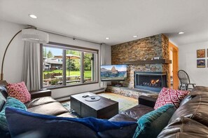 Uma sala de estar moderna com lareira de pedra, assentos confortáveis, uma TV de tela plana e vista para um jardim através de grandes janelas.