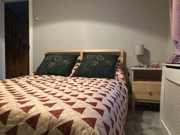 Una cama con una colcha de diseño geométrico, dos almohadas verde oscuro con diseños de hojas, un cabecero de madera, una mesita de noche con una lámpara y un pequeño estante con un tarro.