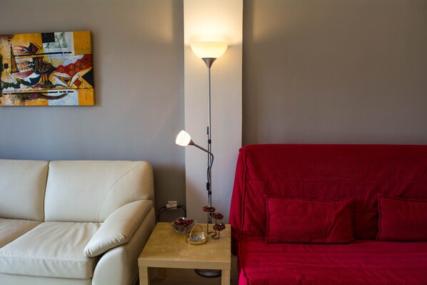 A room with a beige sofa, a red sofa, a small wooden table with a lamp, and a painting on the wall.