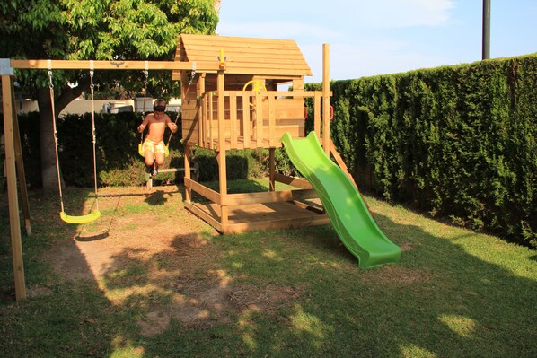 Une structure de jeu en bois, avec un toboggan et des balançoires, installée dans un jardin.