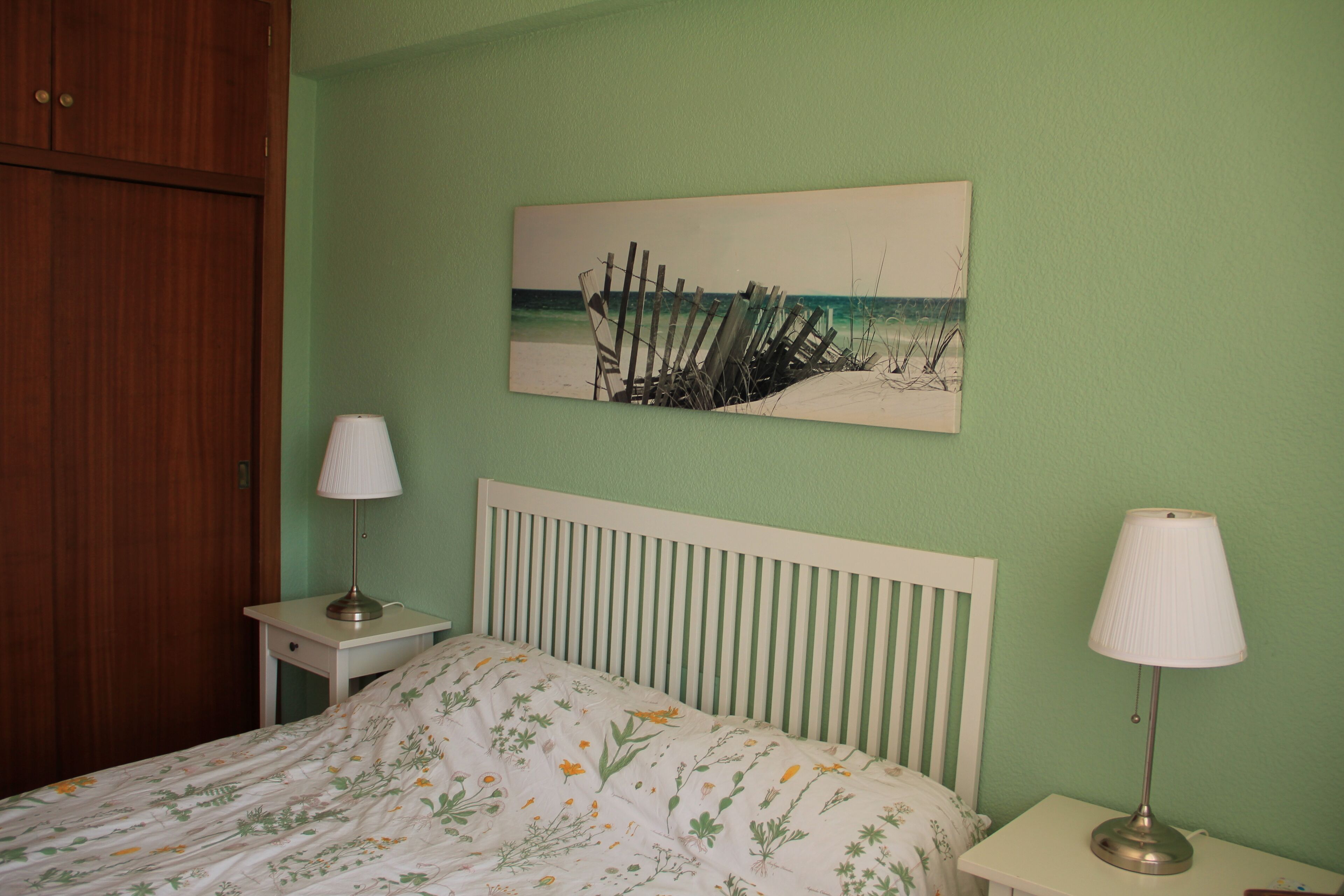 A bedroom with a bed, two bedside tables, a nightstand, a wooden wardrobe, and a beach-themed artwork on the wall.
