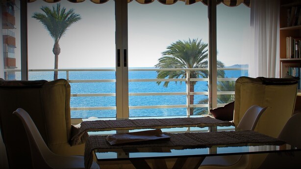 A room with a view of the sea, a table with a woven table runner, and a bookshelf. - seafront apartment with panoramic view to the ocean (El Campello, Alicante)