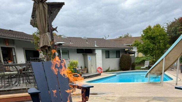 Ein Beton-Feuerkorb mit brennenden Holzscheiten, ein Pool, Gartenmöbel und ein Haus im Hintergrund.