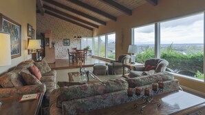 Ein geräumiges Wohnzimmer mit hoher Decke, großen Fenstern mit Blick auf Grünflächen und eine entfernte Stadt sowie ein Essbereich mit Holztisch und Stühlen.